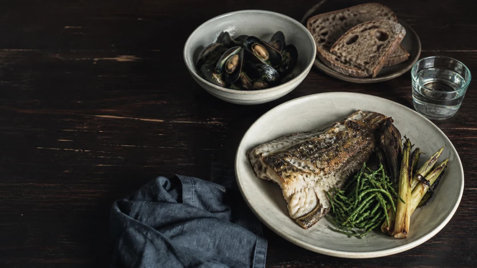 Table set with grilled fish, greens, mussels, bread, and water