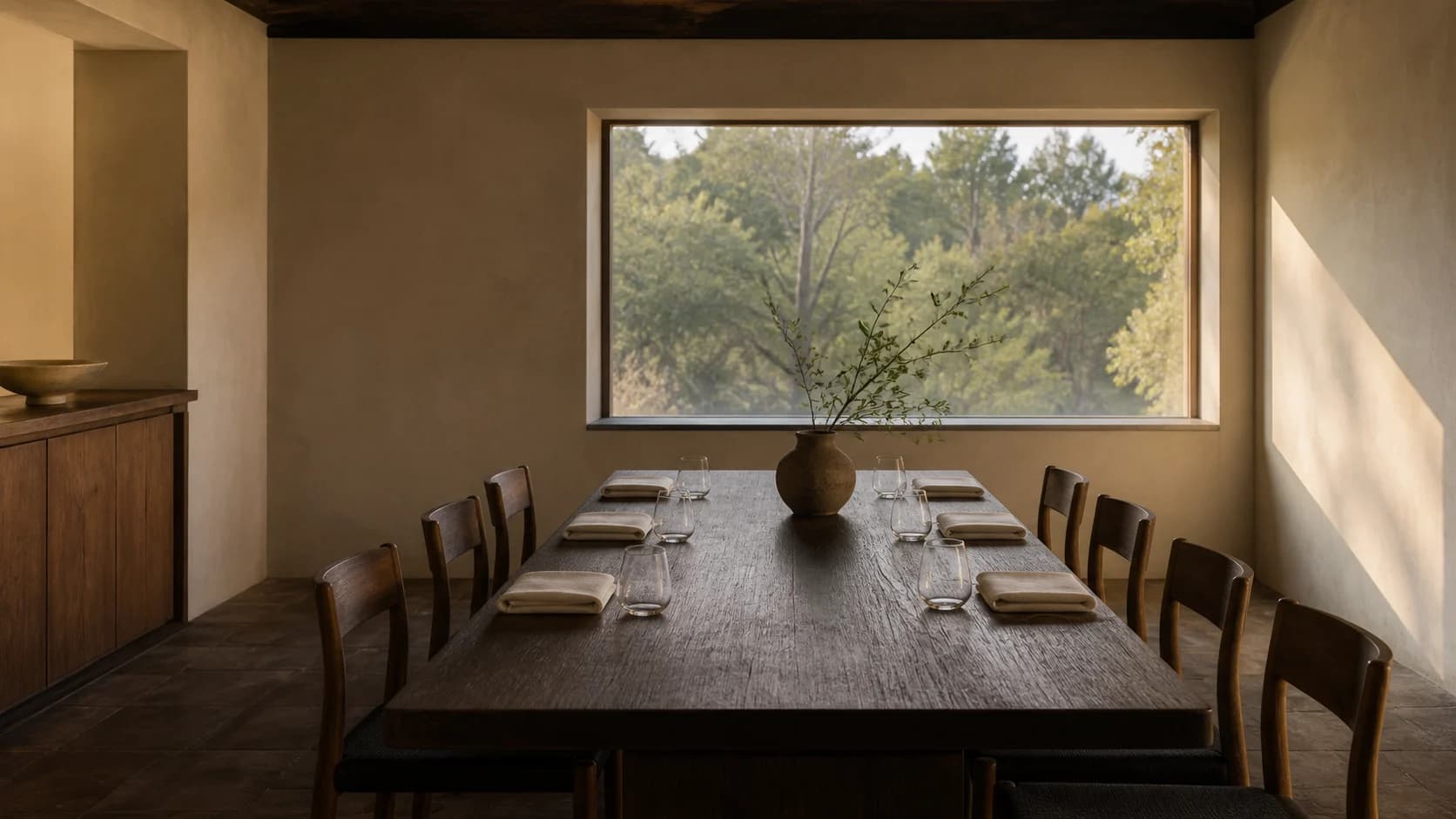 An intimate dining room with a long dark wood table, linen place settings and a garden view